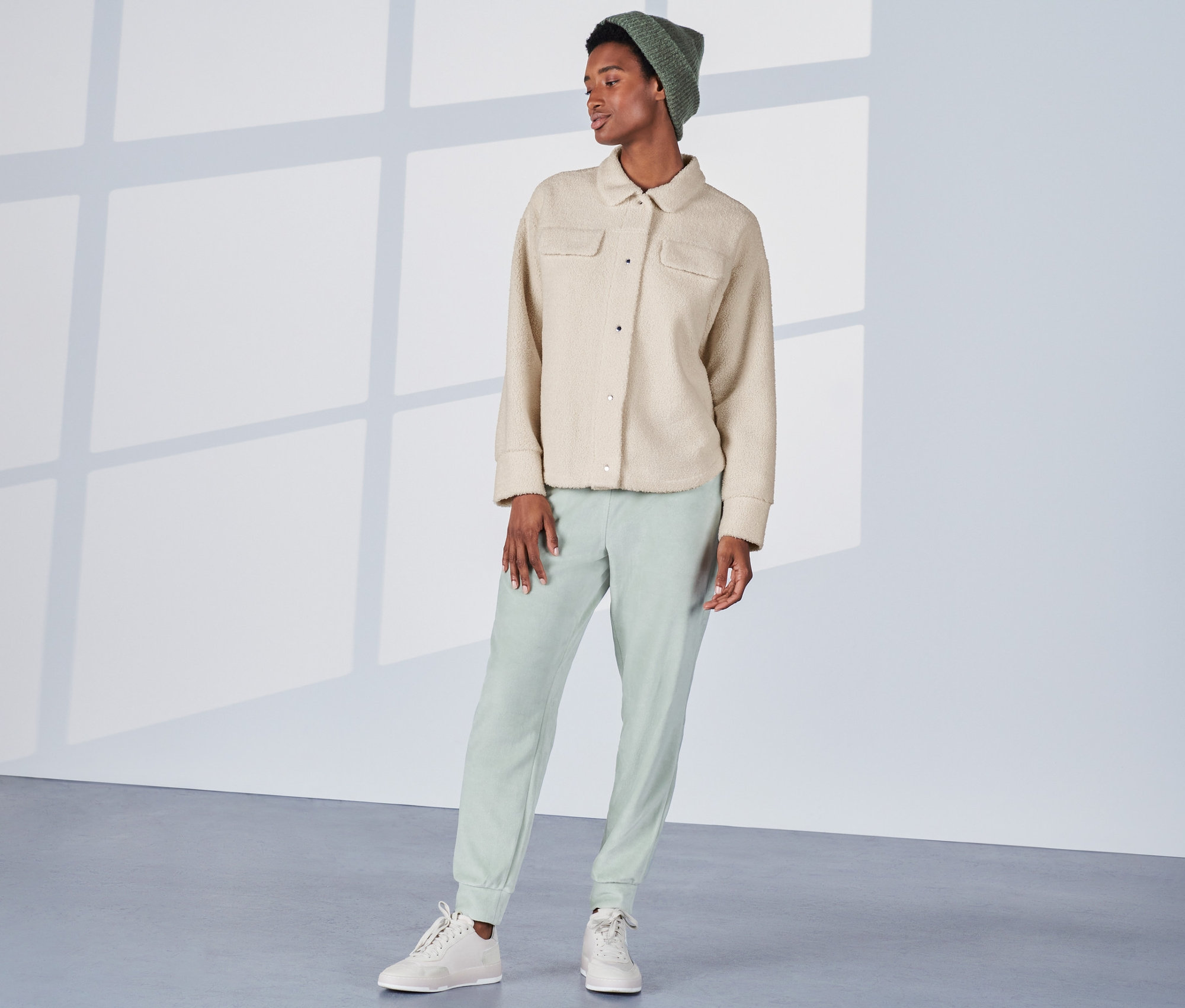 Frau steht und trägt ein cremeweißes Teddy-Overshirt, grüne Hosen und Mütze.