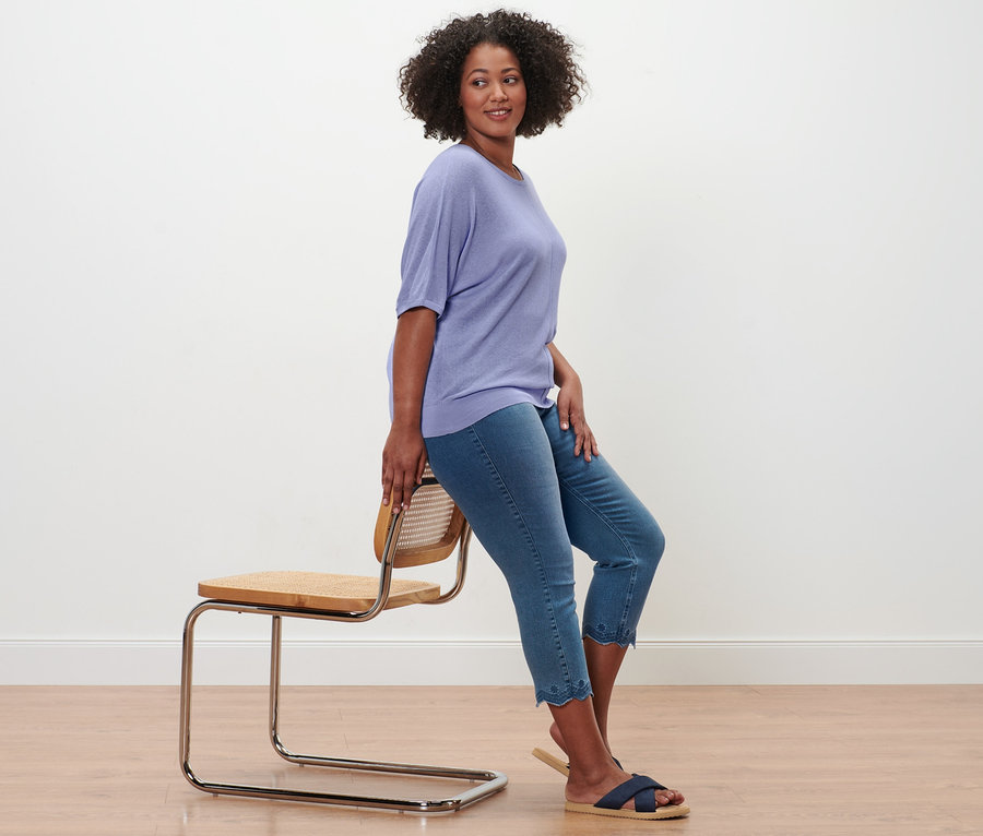 Eine Frau sitzt seitlich auf einem Stuhl und trägt ein Feinstrick-Shirt aus Leinenmix, 7/8-Treggings mit Lochstickerei und Komfortsandalen.