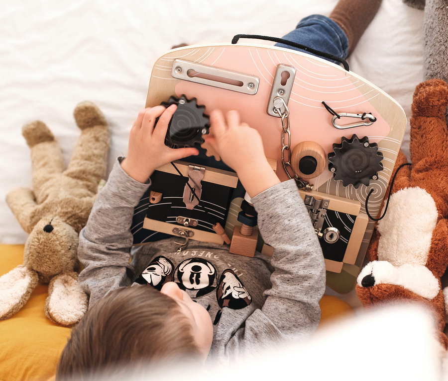 Ein Kind spielt mit dem small foot Motorikbrett »Schlösser und Rotation« auf einem Bett, umgeben von Stofftieren.