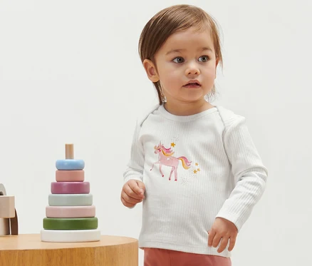 Kleinkind trägt ein geripptes Shirt mit Einhorn. Daneben steht ein Holzspielzeug.