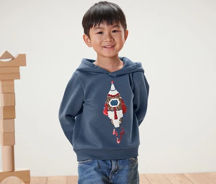 Ein lächelnder Junge posiert in einem Kinder-Sweatshirt mit Wendepailletten, Rakete oder Print (Gr. 86/92).