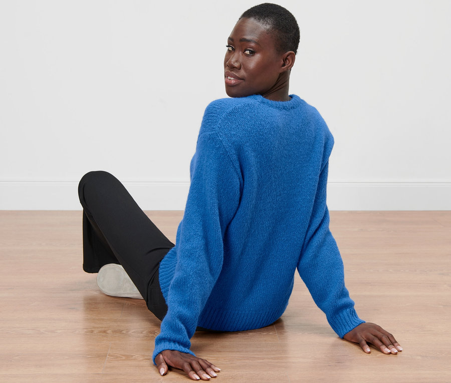 Eine Frau sitzt auf dem Boden und blickt über ihre Schulter, sie trägt einen kobaltblauen Grobstrickpullover mit Wolle.