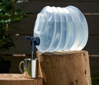 Ein faltbarer Wasserkanister, ca. 10 l, steht auf einem Holzscheit und befüllt einen silbernen Becher mit Wasser.