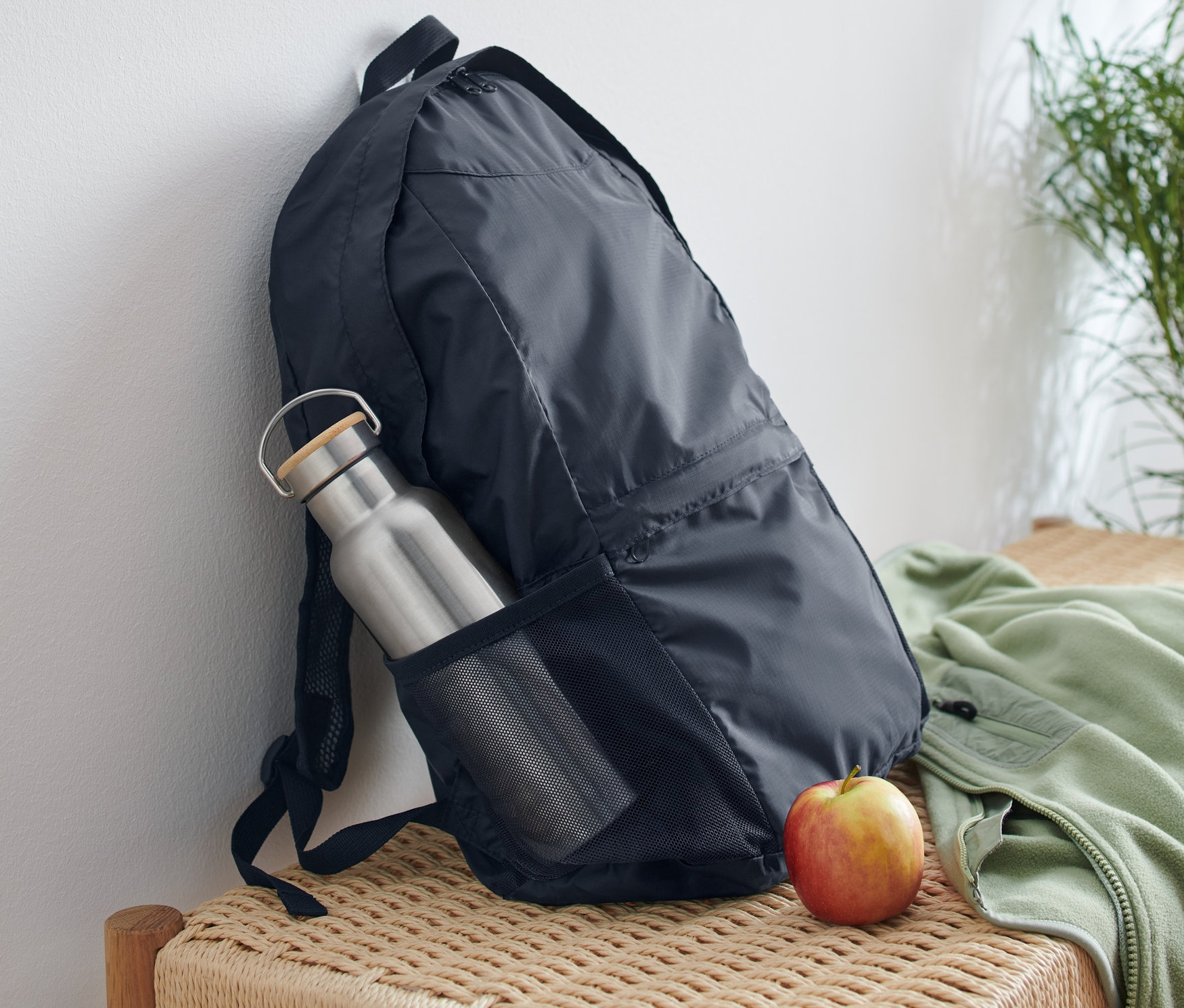 Ein faltbarer Rucksack lehnt an einer weißen Wand, mit einer Wasserflasche in der Seitentasche und einem Apfel daneben auf einer geflochtenen Bank.