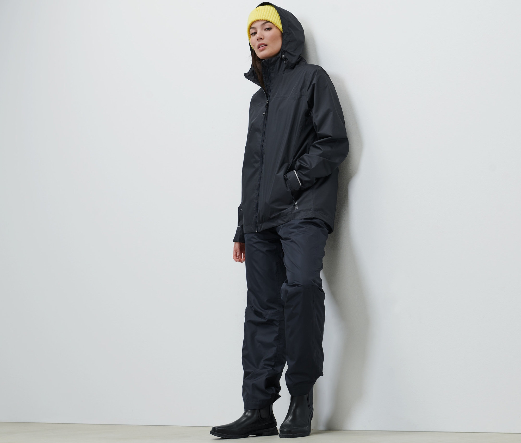 Frau trägt eine dunkelblaue Unisex Regenjacke, Hose und eine gelbe Mütze.
