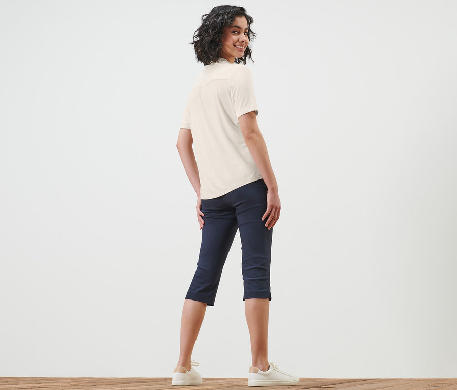 Frau blickt über die Schulter und trägt ein weißes Blusenshirt mit Knopfleiste, dunkelblaue Caprihosen und weiße Turnschuhe.