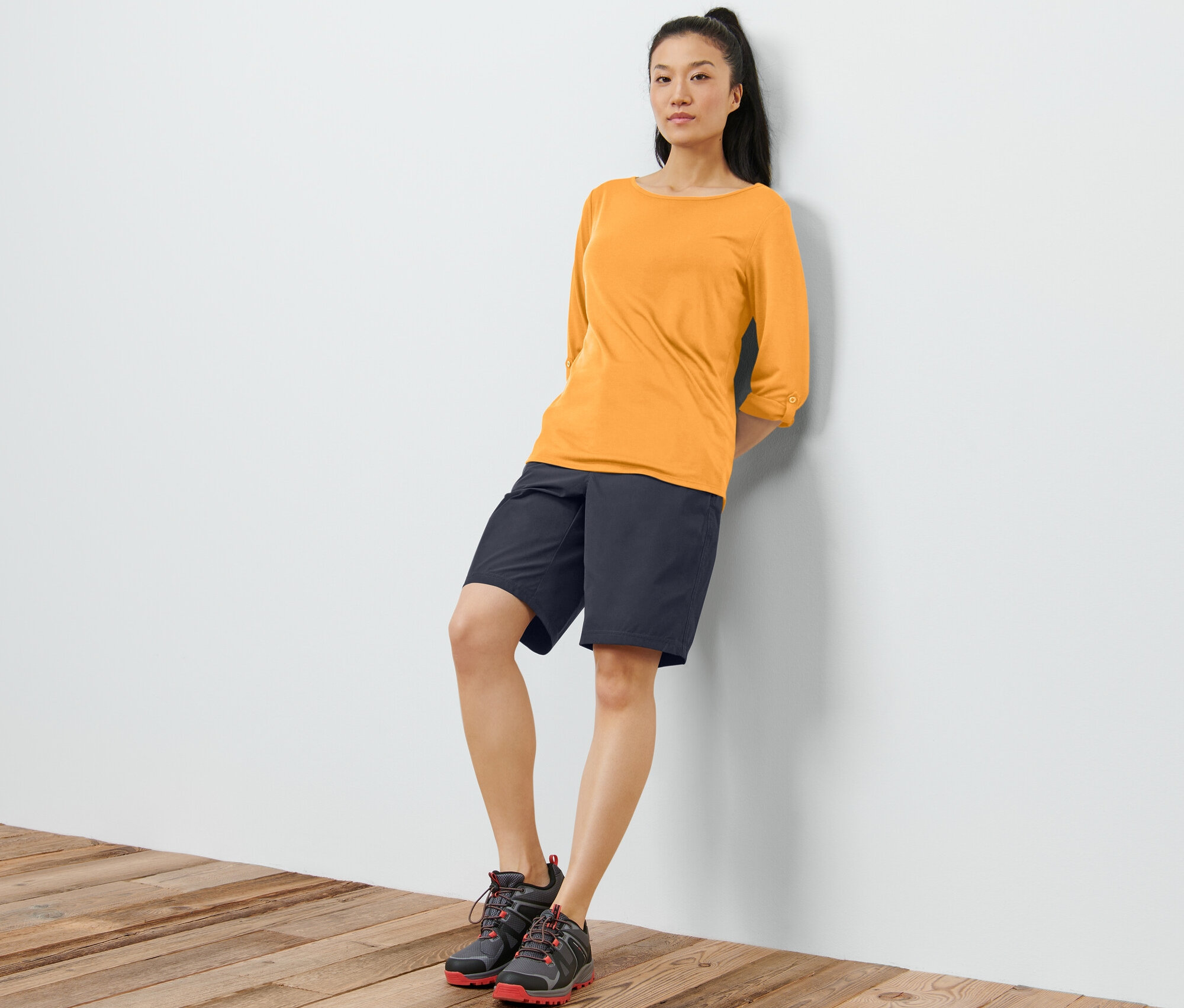 Eine Frau lehnt an einer Wand und trägt ein oranges Funktionsshirt mit 3/4-Ärmeln, schwarze Shorts und Schuhe.