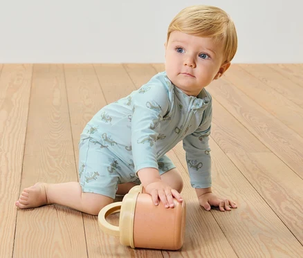 Ein Baby krabbelt auf einem Holzboden und trägt einen blauen Baby-Einteiler mit UV-Schutzfaktor 80, Drache, während es mit einem Becher spielt.