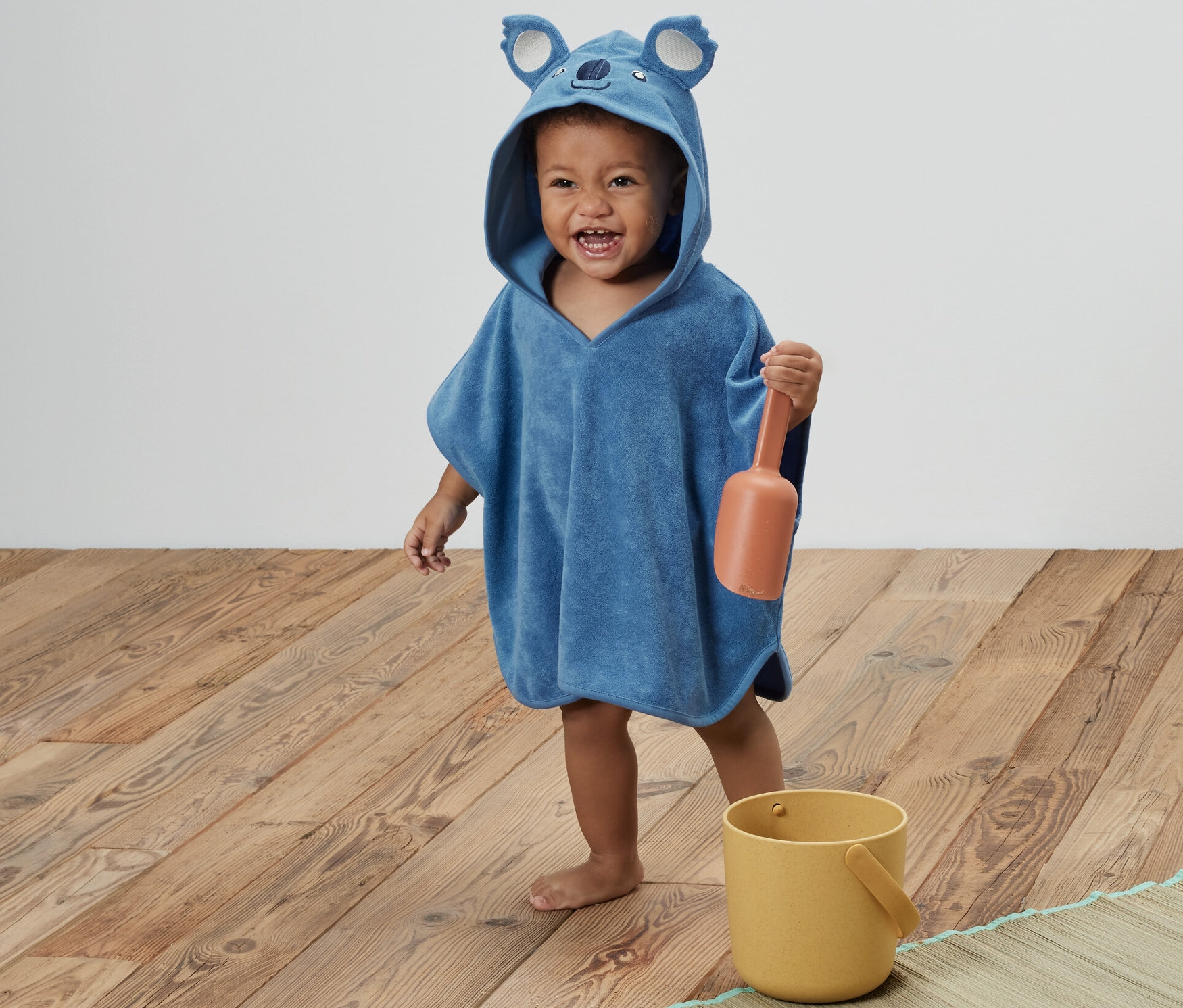 Ein Kind trägt einen blauen Kinder-Frottierponcho mit Koala-Kapuze und hält eine orangefarbene Schaufel. Daneben steht ein gelber Eimer.