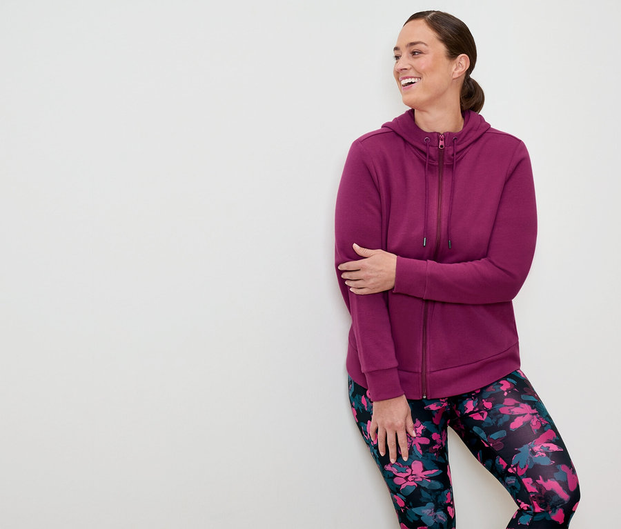 Eine Frau in einem violetten Kapuzenpullover mit Reißverschluss und floralen Leggings lehnt an einer weißen Wand und lächelt.