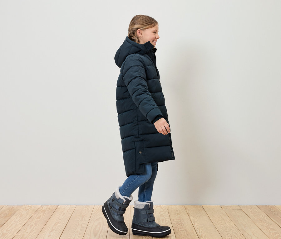 Mädchen in blauem Steppmantel und Schneestiefeln geht über Holzboden.