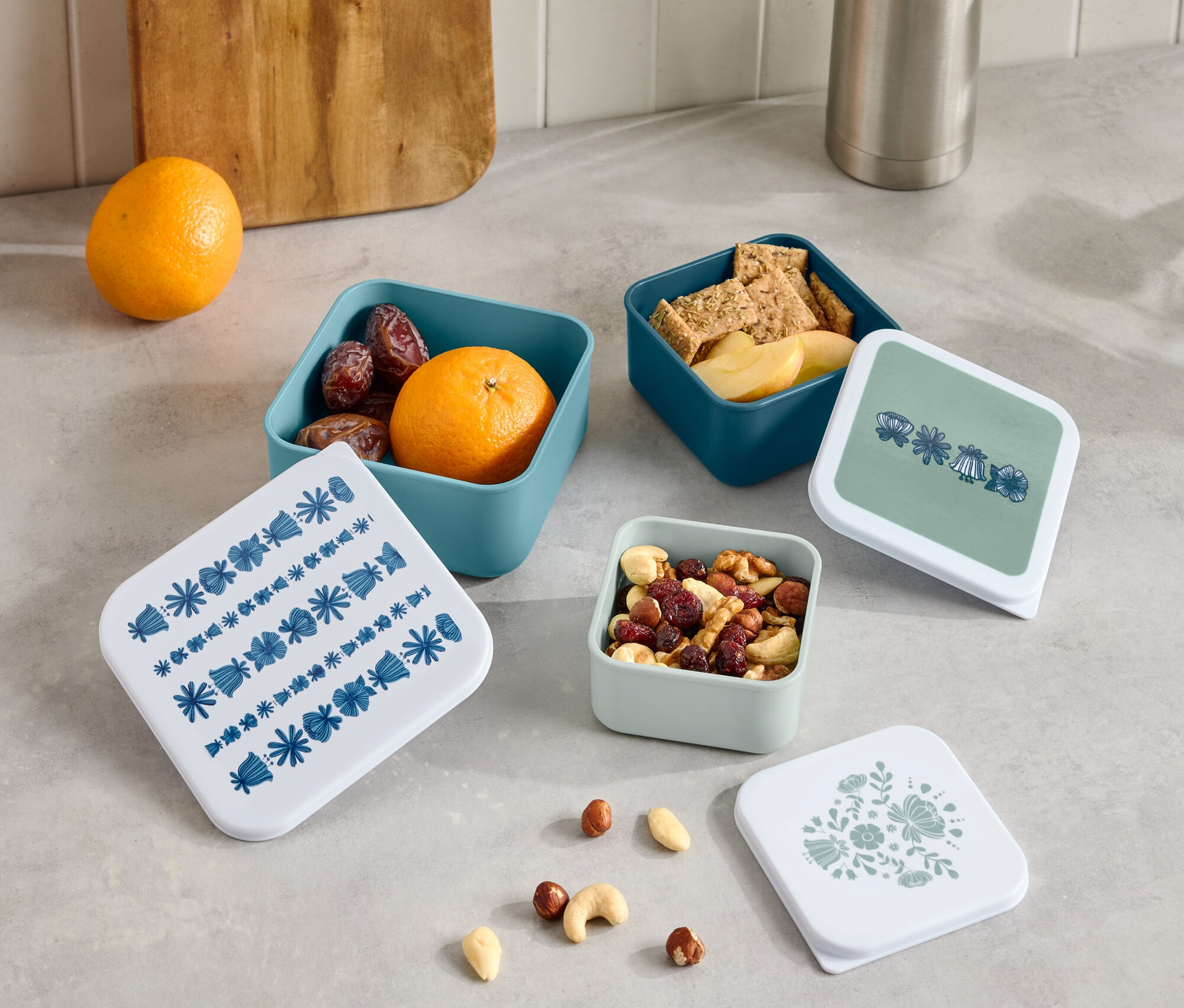 Auf einer Theke stehen eine Thermoskanne und ein Holzbrett neben vier kleinen, offenen Schachteln mit Obst, Crackern und Nüssen sowie vier Deckeln mit verschiedenen Mustern.