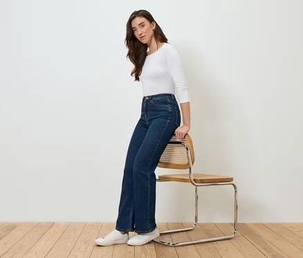 Frau sitzt seitlich auf einem Stuhl, trägt blaue Jeans und ein weißes T-Shirt.