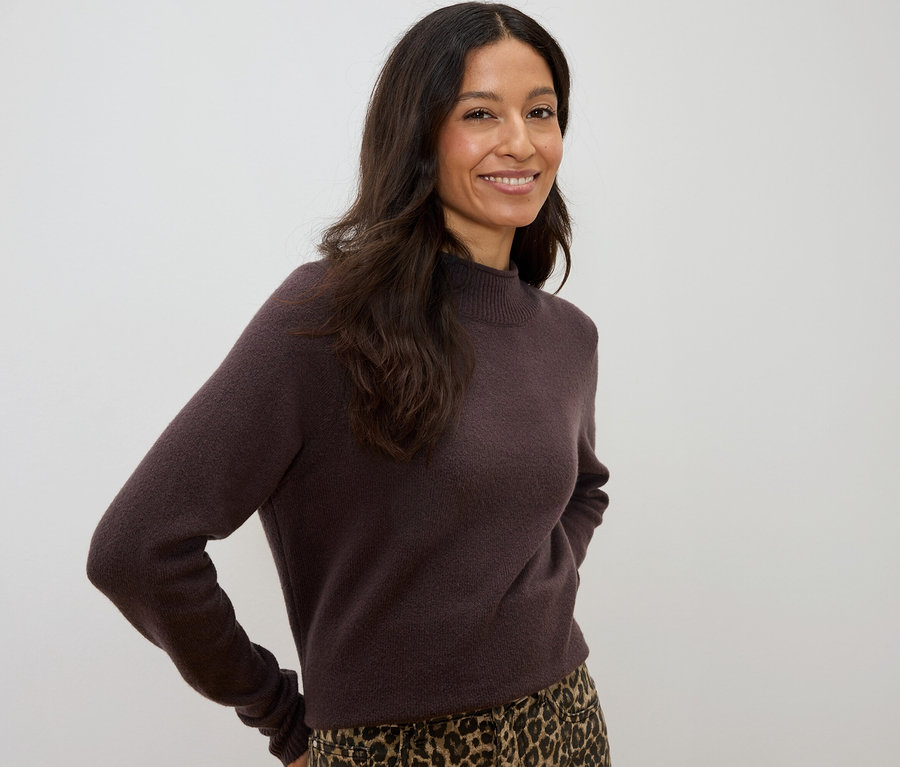 Frau mit dunklen, gewellten Haaren, die einen braunen Pullover und einen Rock mit Leopardenmuster trägt und lächelt.