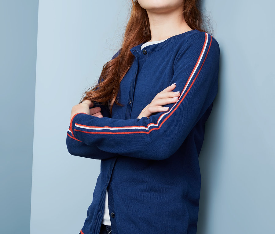 Mädchen mit braunen Haaren posiert in Strickjacke mit blauen Streifen an den Ärmeln.