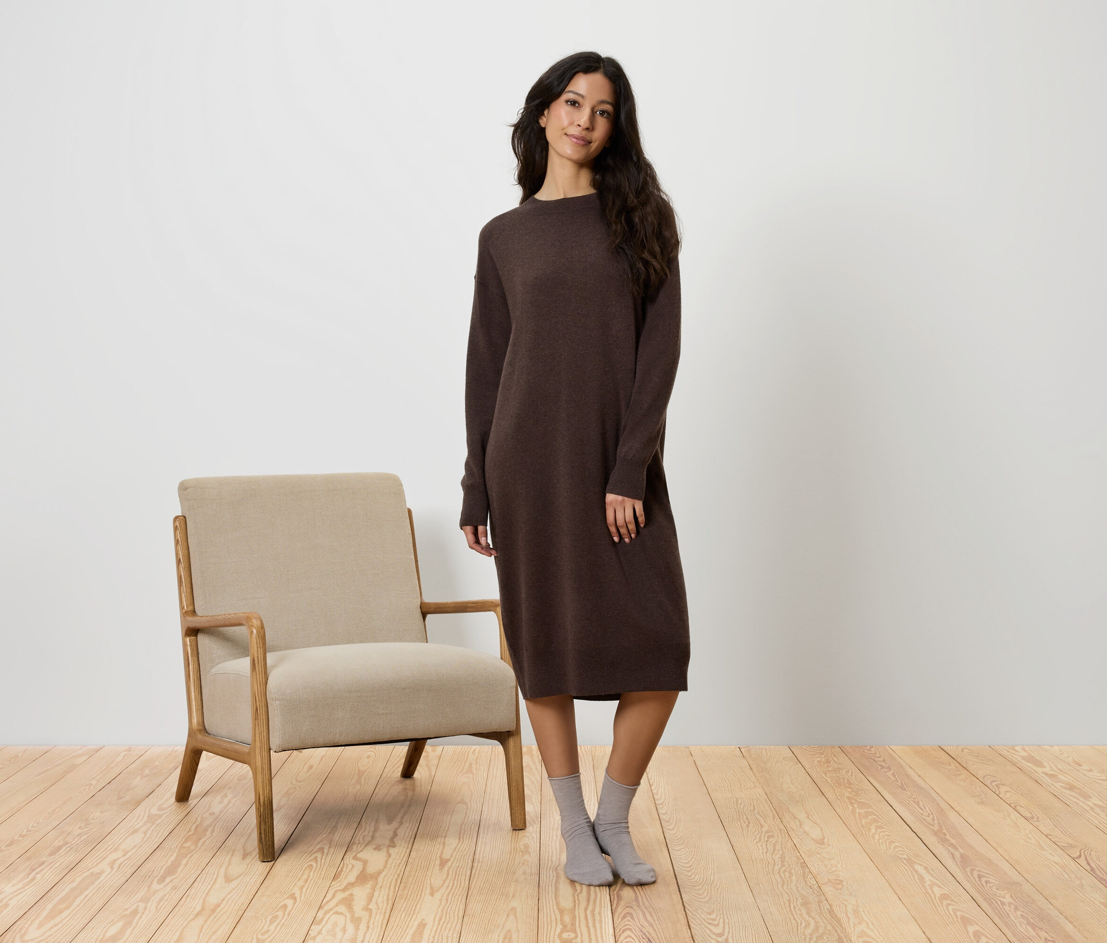 Eine Frau steht neben einem beigen Sessel und trägt ein braunes Kleid und graue Socken.