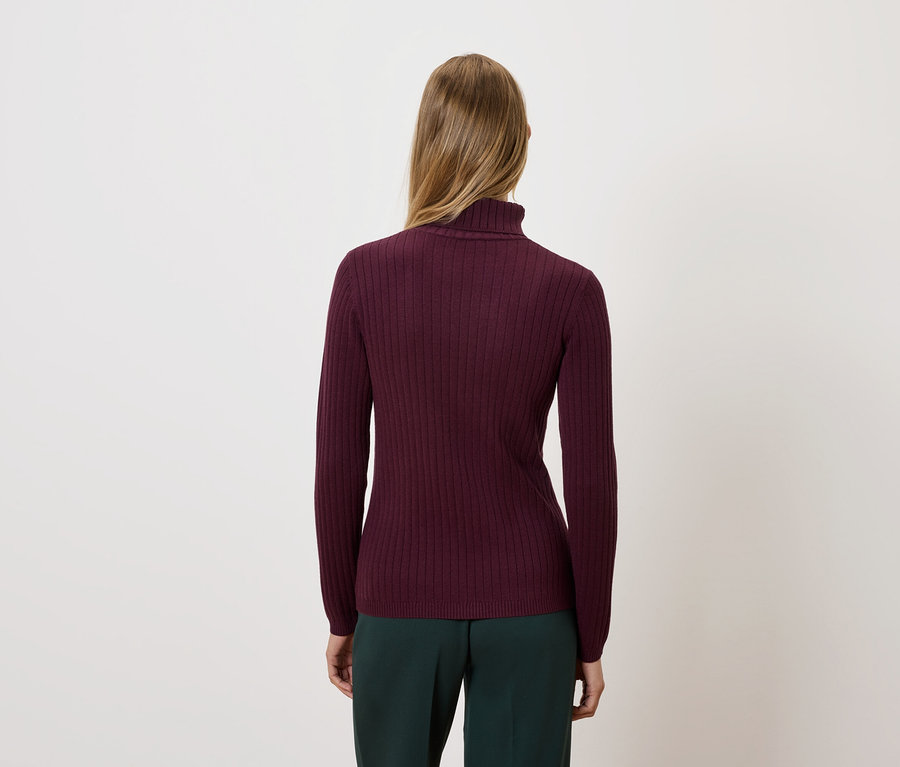 Frau mit dem Rücken zur Kamera trägt einen bordeauxfarbenen Rollkragenpullover und dunkelgrüne Hosen.