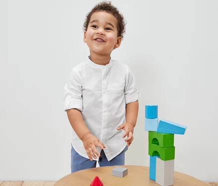 Ein Junge in einem Kinder-Hemd spielt mit bunten Holzbausteinen auf einem Tisch.
