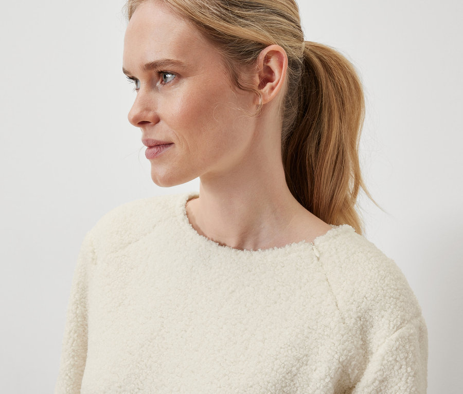 Detailaufnahme des Profils einer blonden Frau mit zum Pferdeschwanz hochgebundenen Haaren, die einen cremefarbenen Pullover trägt.