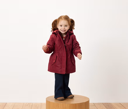 Ein Mädchen mit roten Zöpfen steht auf einem Holzsockel und trägt eine burgunderrote Jacke und dunkelblaue Hosen.