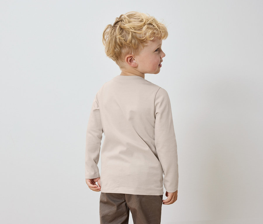 Ein Junge mit blonden Haaren steht mit dem Rücken zur Kamera, er trägt ein beiges Langarmshirt und braune Hosen.