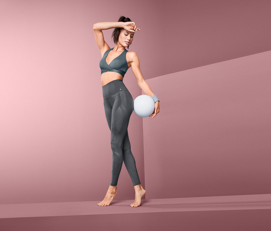 Frau posiert in Bodyforming-Sporttight und Sport-BH, hält einen weißen Ball.