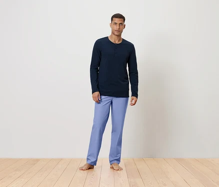 Mann steht barfuß auf einem Holzboden und trägt ein dunkelblaues Langarmshirt und hellblaue Hosen.