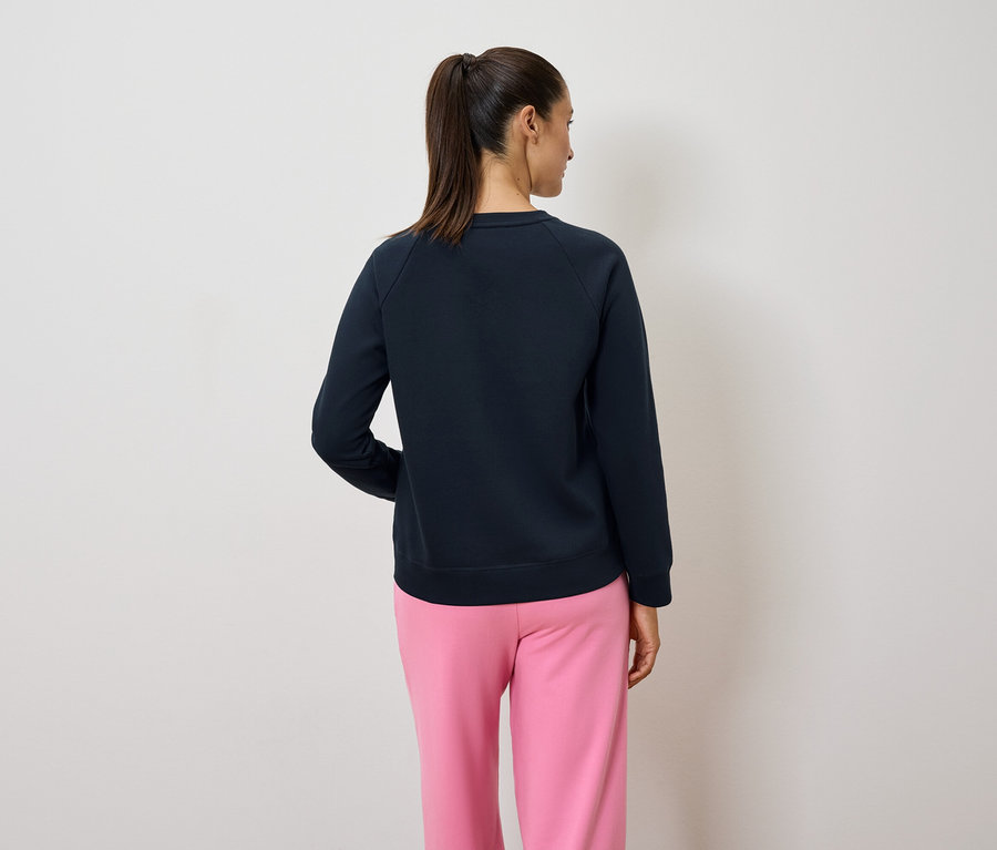 Rückansicht einer Frau mit Pferdeschwanz, die einen dunkelblauen Pullover und eine rosafarbene Hose trägt.