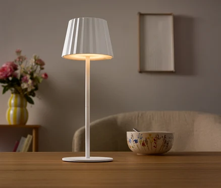 Auf einem Tisch steht eine wei�e Lampe mit einem plissierten Lampenschirm. Im Hintergrund befinden sich eine Blumenvase und eine Sch�ssel.