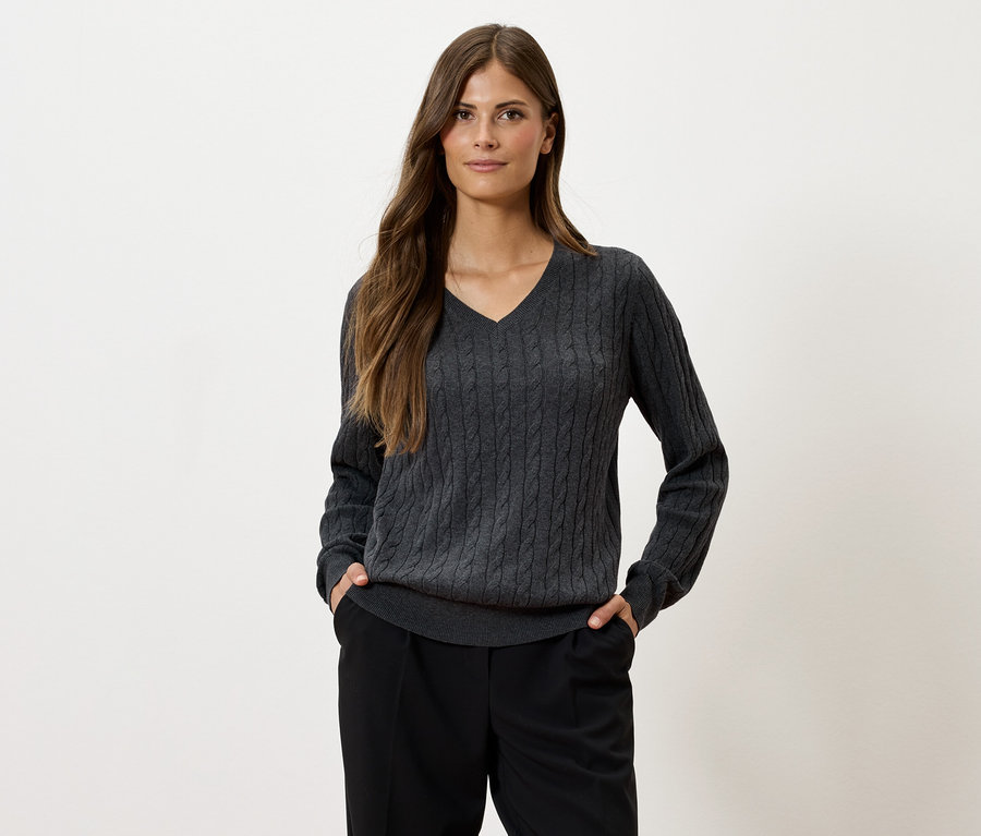 Eine Frau mit langen braunen Haaren posiert vor einem hellen Hintergrund und trägt einen grauen Zopfstrickpullover mit V-Ausschnitt und eine schwarze Hose.