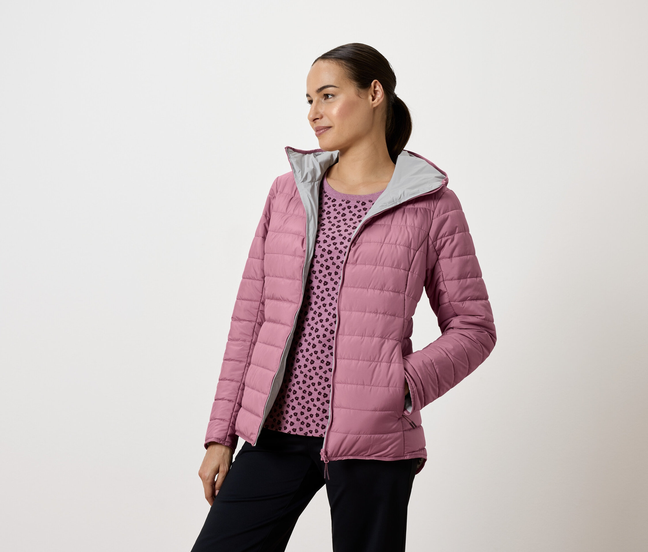 Frau trägt eine rosa Steppjacke mit Kapuze und marineblaue Hosen, sie schaut nach links.