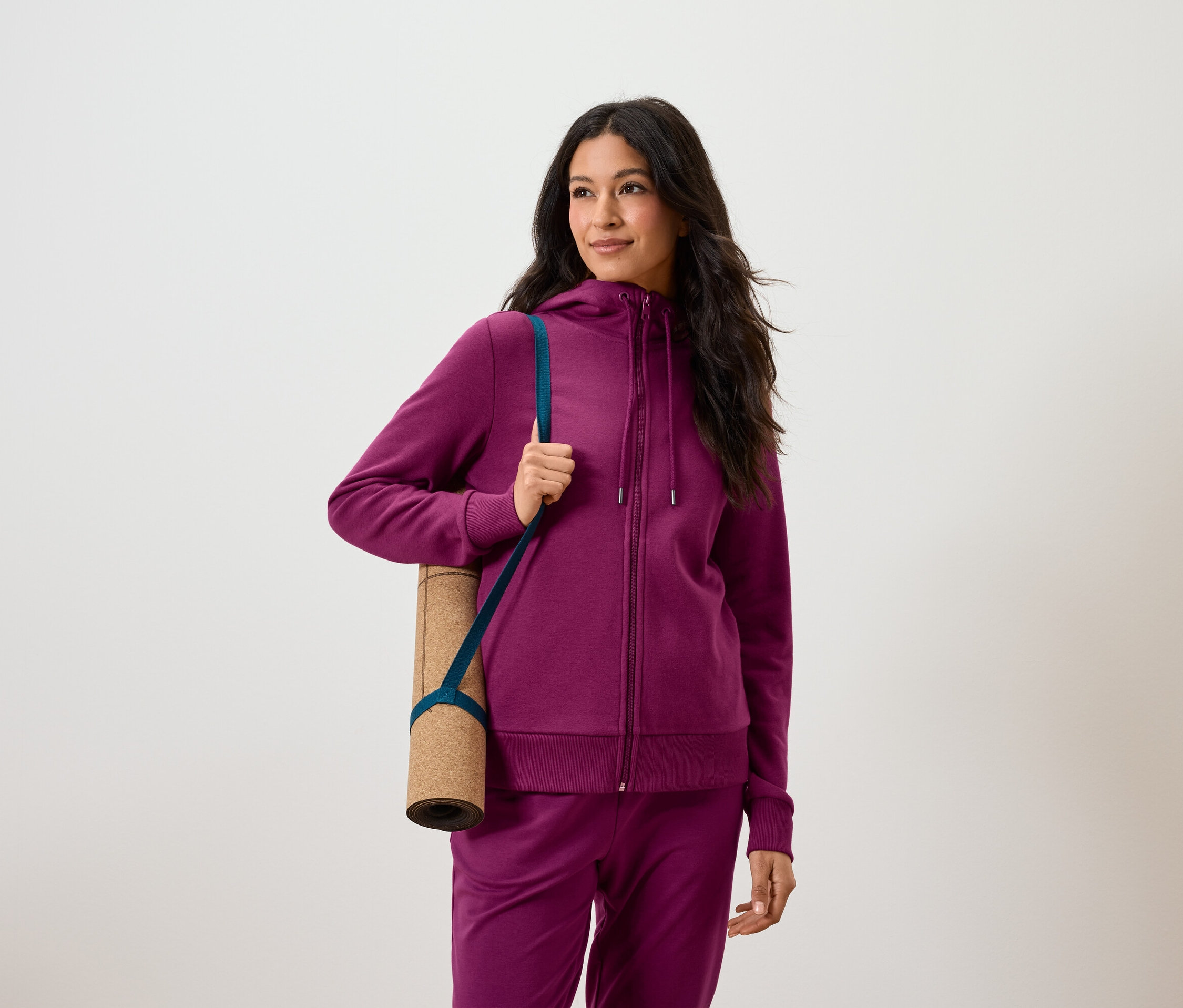 Frau mit dunklen Haaren in lila Trainingsanzug hält eine zusammengerollte Yogamatte unter dem Arm.