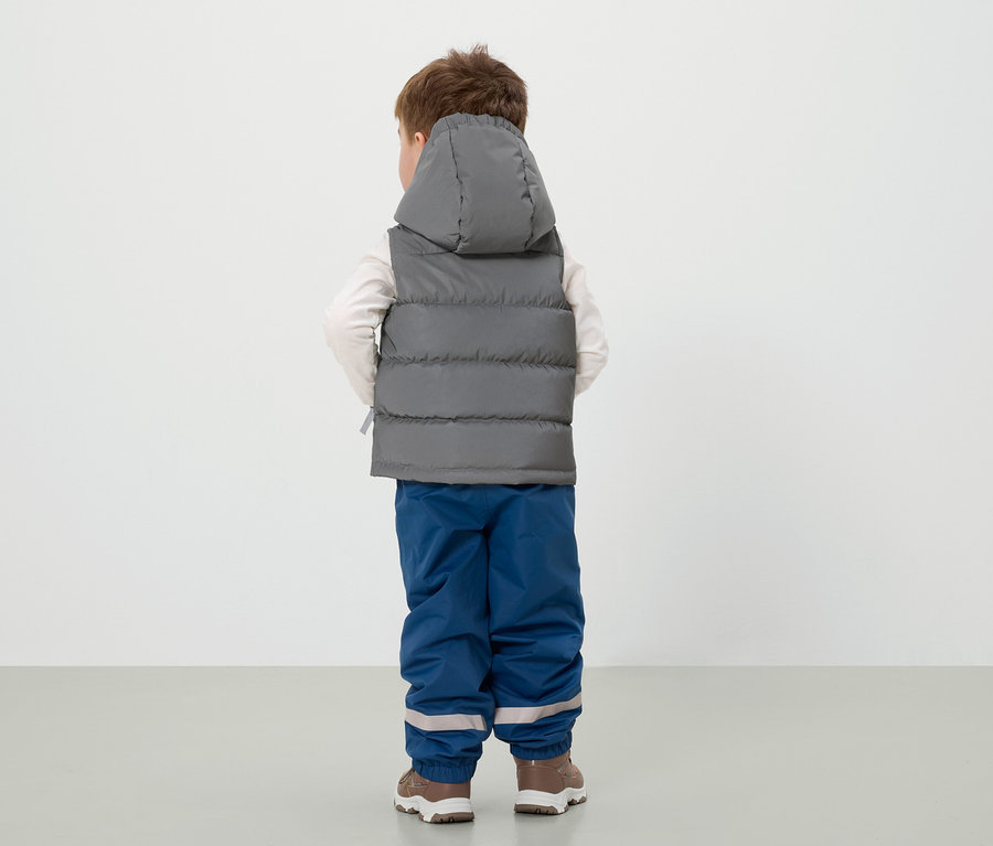 Junge steht mit dem Rücken zur Kamera, trägt eine graue Weste mit Kapuze, blaue Hosen und braune Schuhe.