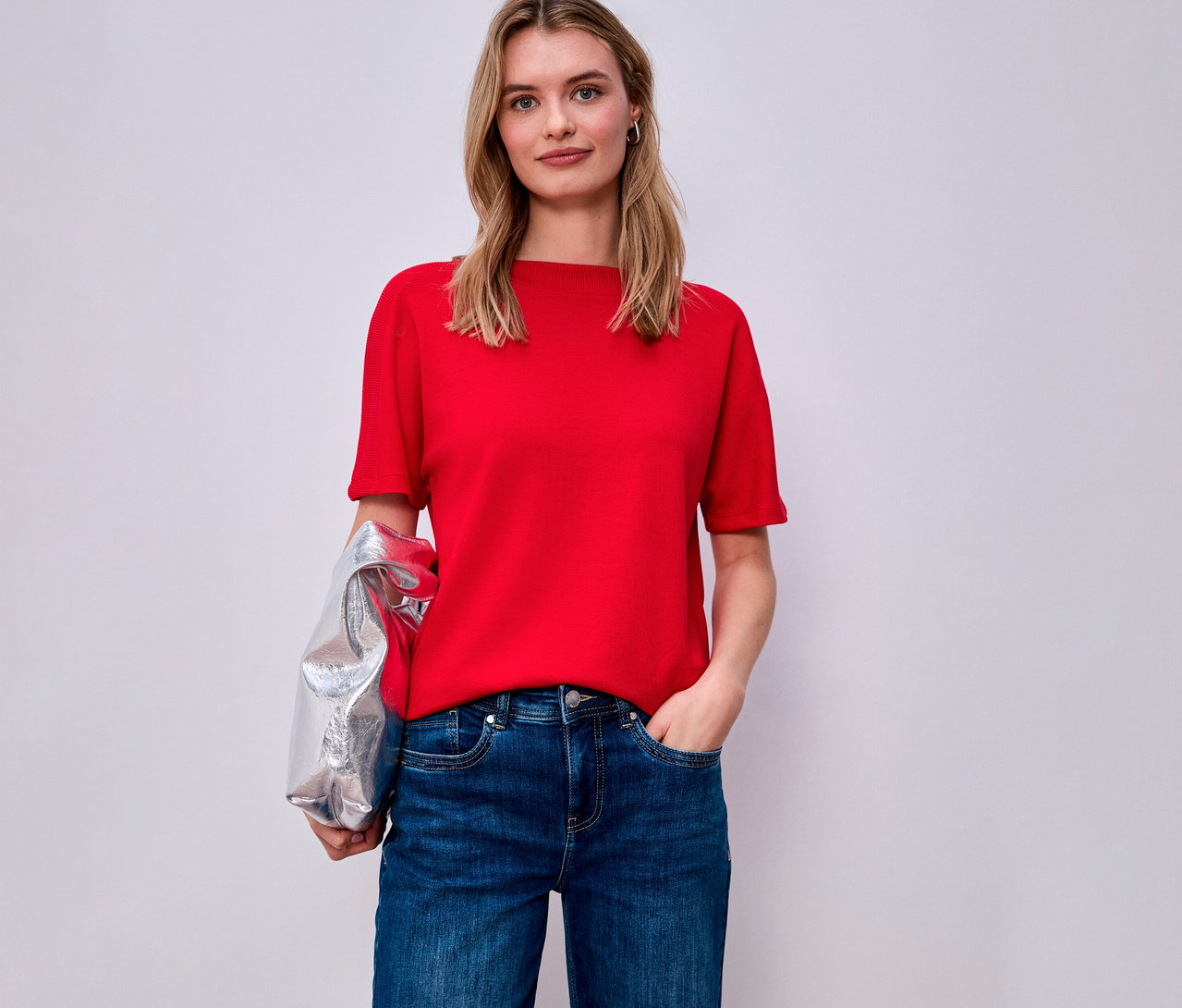 Frau mit rotem Street One Kurzarmshirt und Jeans, silberne Jacke über dem Arm.