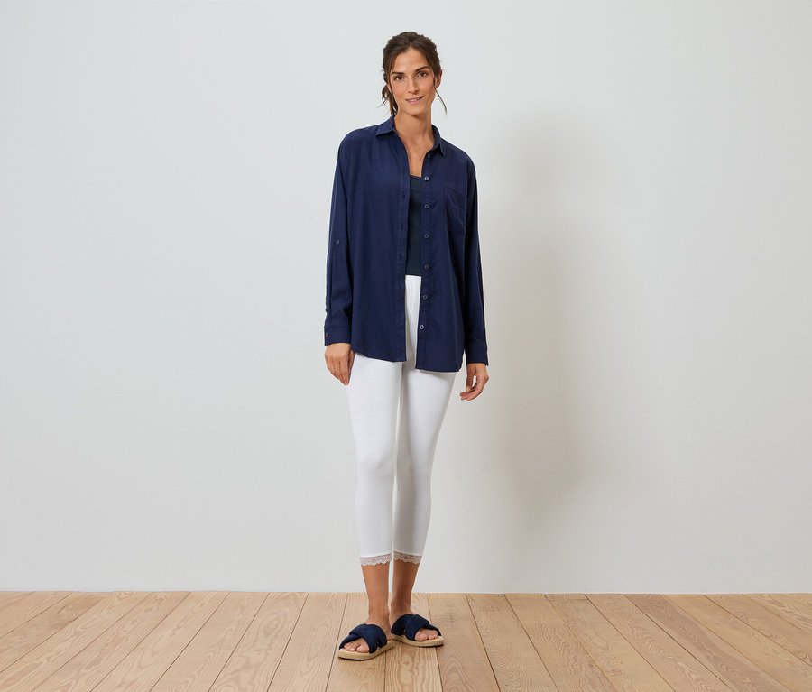 Frau steht vor einer weißen Wand und trägt ein blaues Hemd, weiße Leggings und blaue Hausschuhe.