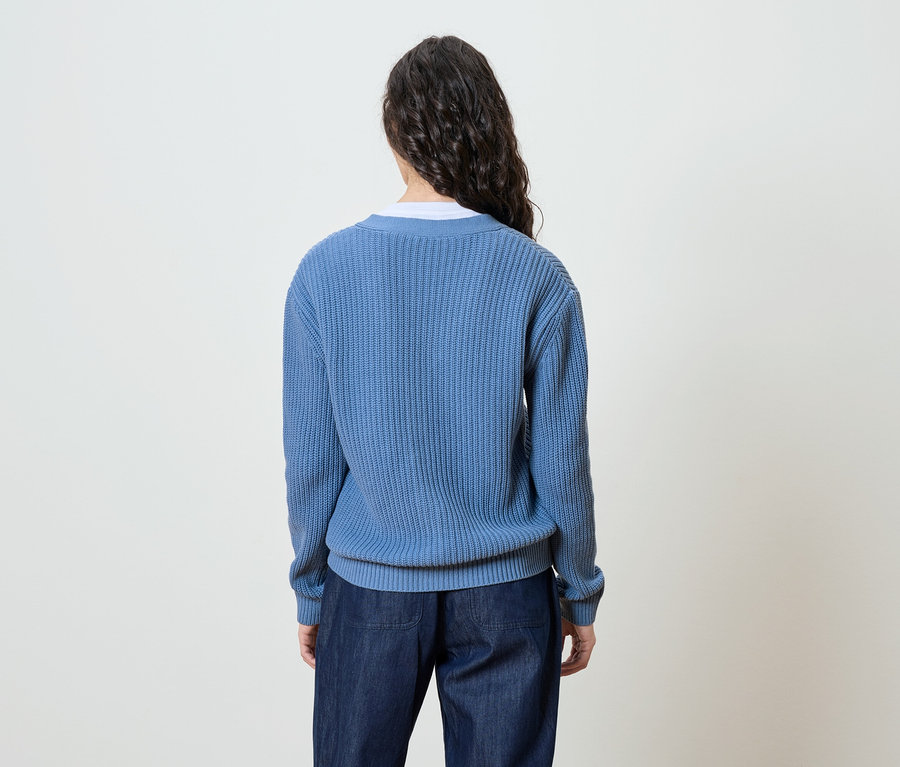 Eine Frau mit langen, dunklen, gewellten Haaren steht mit dem Rücken zur Kamera und trägt einen blauen Strickpullover und Jeans.