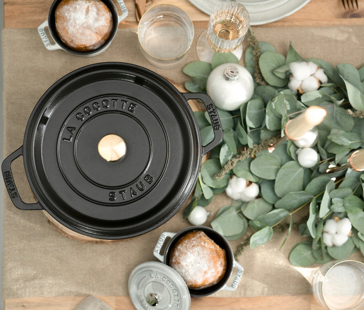 Auf einem Tisch stehen mehrere schwarze STAUB »Cocotte« Töpfe (24 cm, 3,7 l; 26 cm, 5,2 l; 24 cm, 6,7 l) neben Gläsern und Dekoration.