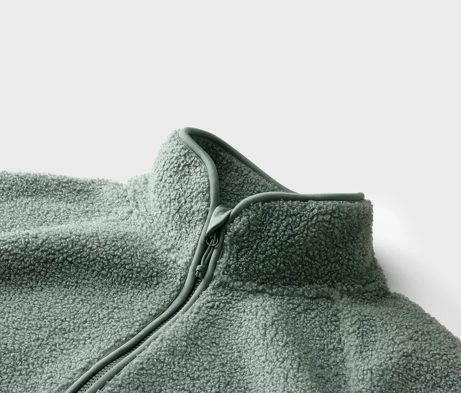 Nahaufnahme einer hellgrünen Fleecejacke mit Reißverschluss und hohem Kragen vor einem weißen Hintergrund.