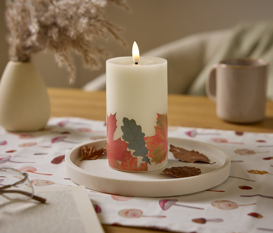Eine brennende, weiße Kerze mit Blättermuster steht auf einem Tisch mit einem beige farbenen Krug und einer Vase.