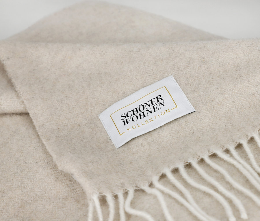 Nahaufnahme einer gefalteten, hellbeigen Decke mit weicher Textur und weißen Fransen. Ein weißes Etikett mit goldener Schrift "SCHÖNER WOHNEN KOLLEKTION" ist auf die Decke genäht.