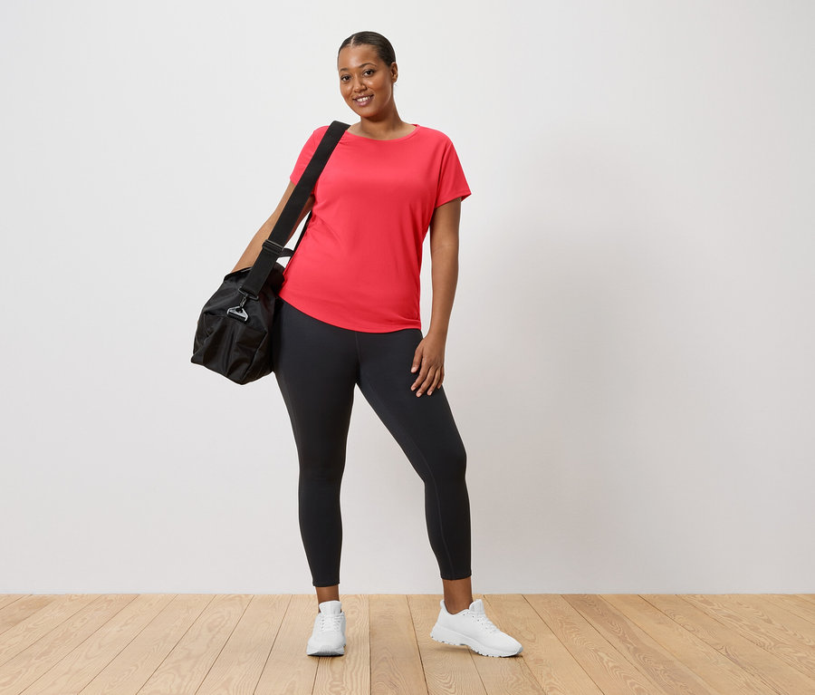 Frau in rotem T-Shirt und Leggings mit schwarzer Sporttasche über der Schulter und weißen Turnschuhen.