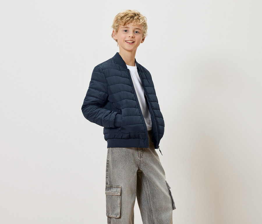 Ein blonder, lächelnder Teenager trägt eine marineblaue Steppjacke und graue Cargohosen.