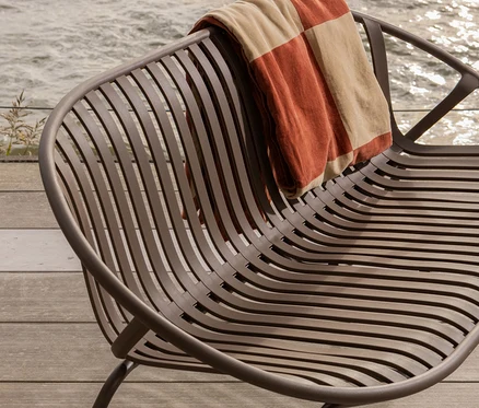 Ein brauner Metallstuhl mit einer gestreiften Decke steht auf einer Holzterrasse am Wasser.
