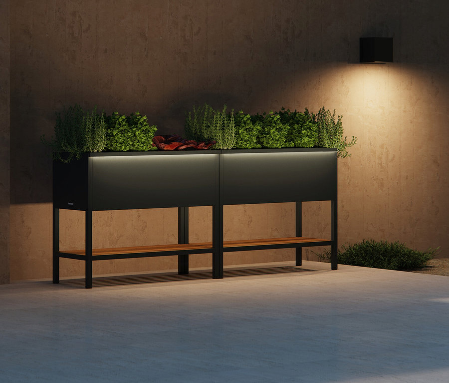 Zwei schwarze Pflanzkästen mit Holzablage stehen vor einer Wand, beleuchtet von einer Wandlampe.