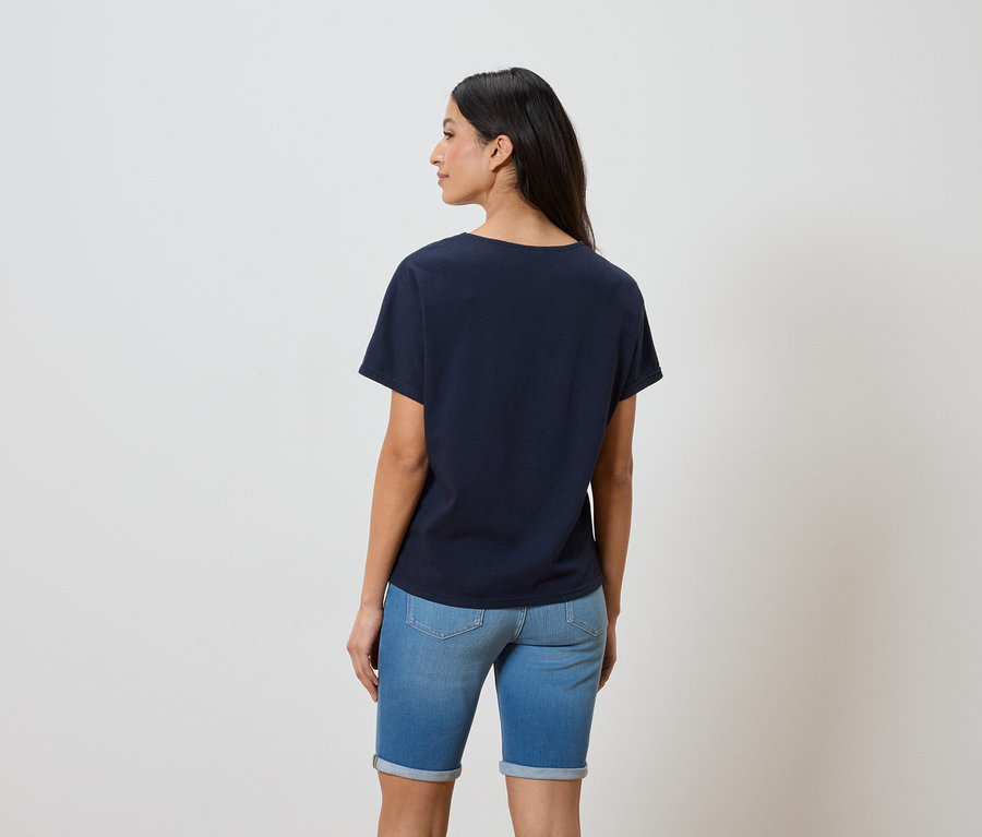 Rückansicht einer Frau mit dunklen Haaren, die ein marineblaues T-Shirt und Jeansshorts trägt.