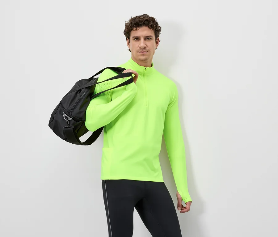 Mann mit schwarzer Sporttasche über der Schulter, trägt ein neongelbes Langarmshirt und schwarze Leggings vor einem weißen Hintergrund.