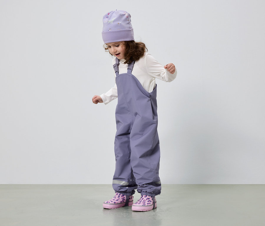 Ein kleines Mädchen mit lockigem Haar trägt eine lila Mütze, eine weiße Bluse, eine lila Hose und rosa Stiefel.
