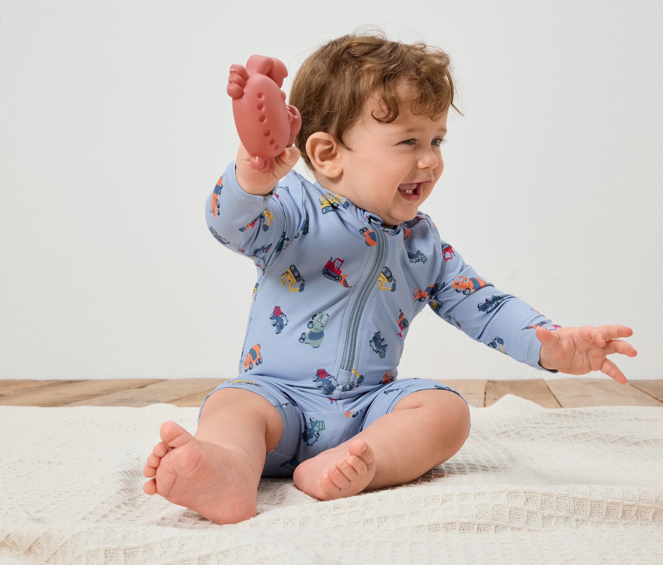 Lachender Säugling mit lockigem Haar, der auf einer weißen Decke sitzt und einen roten Beißring in der Hand hält. Der Säugling trägt einen hellblauen Spielanzug mit bunten Fahrzeugen.