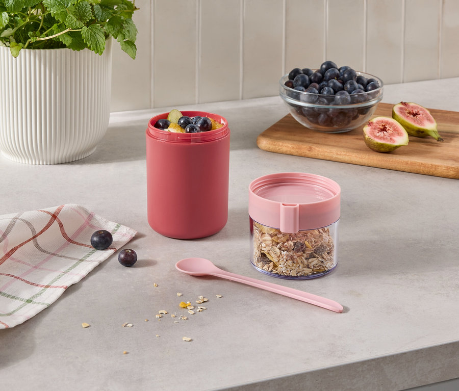 Zwei Behälter auf einer Küchenarbeitsplatte: ein hoher, rosafarbener Behälter mit Früchten und ein durchsichtiger Behälter mit Müsli und rosa Deckel. Daneben liegen ein rosa Löffel, ein kariertes Tuch, Beeren, eine Schale Blaubeeren und halbierte Feigen auf einem Holzbrett.