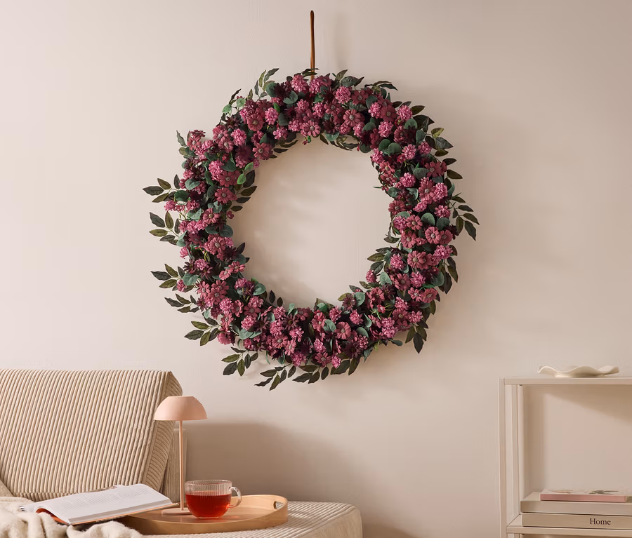Großer Blumenkranz mit lila Blüten und grünen Blättern an einer hellen Wand. Links ein Sofa mit Decke, Buch, rosa Lampe und Teetasse auf einem Tablett. Rechts ein weißes Regal mit Büchern.